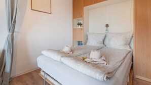 Ein ordentlich bezogenes Bett mit weißer Bettwäsche und Handtüchern, ein Holzschrank und ein kleines Regal mit Pflanze und Büchern.