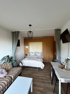 A modern hotel room with a bed, a sofa, a dining table, and a TV.