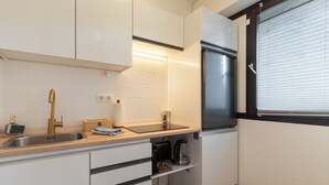 Eine kleine Küche mit weißen Schränken, einem Edelstahlkühlschrank, einem Einbauofen und einem Fenster mit Jalousien.