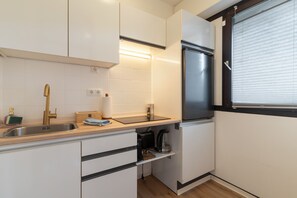 Eine kleine Küche mit weißen Schränken, einem Edelstahlkühlschrank, einem Einbauofen und einem Fenster mit Jalousien.