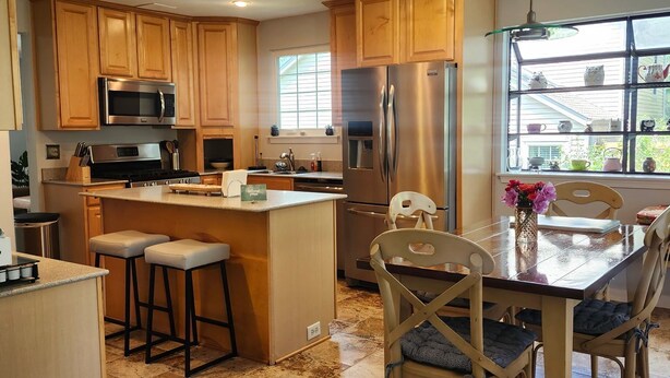 Una cocina moderna con armarios de madera, electrodomésticos de acero inoxidable, una isla central con taburetes y un comedor con mesa y sillas.