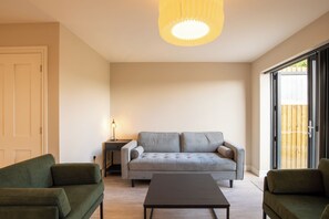 Una sala moderna con un sofá gris, dos sillones verdes, una mesa de centro, una lámpara y un ventanal corredero.