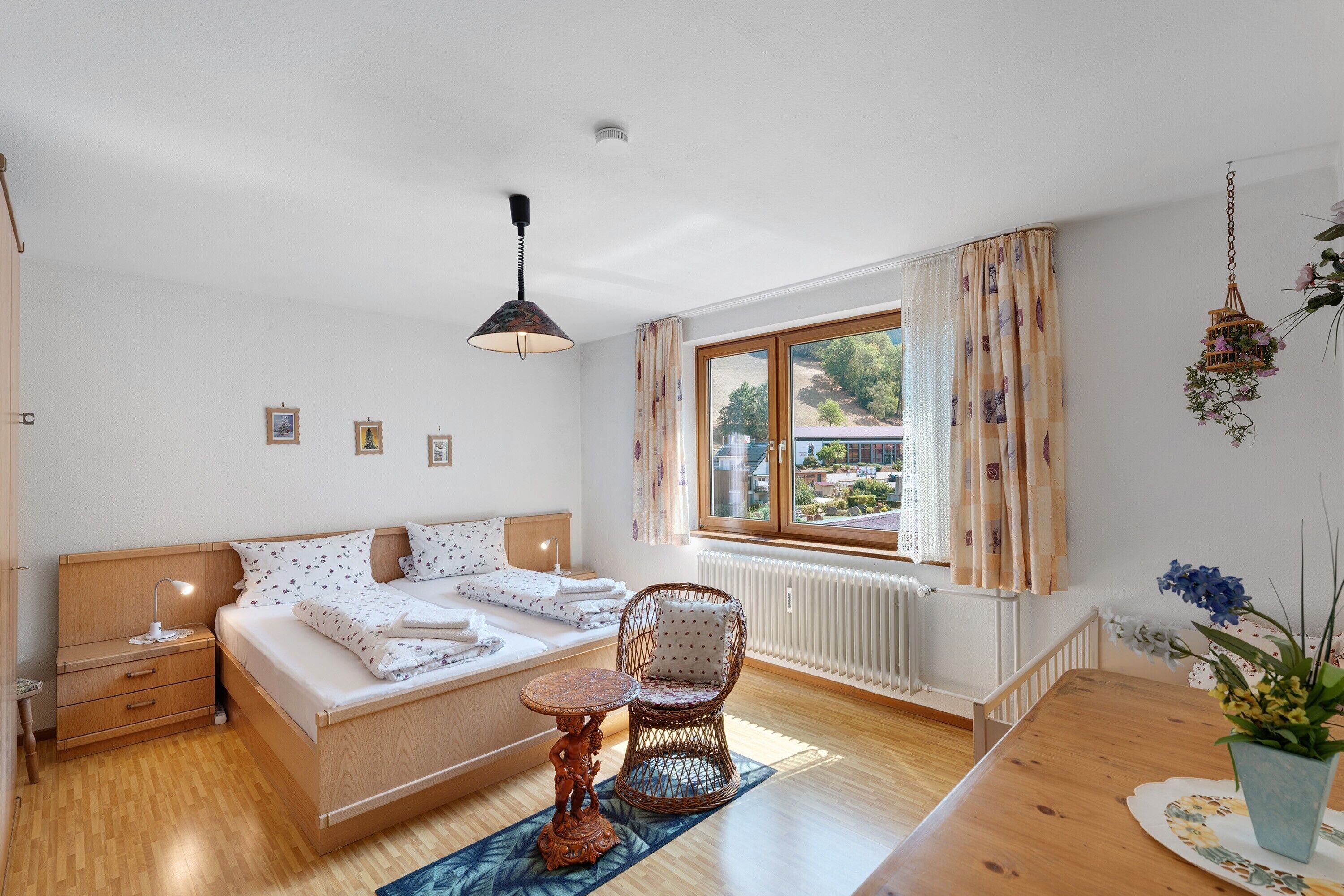 Ein Schlafzimmer mit Bett, Nachttischen, einem Fenster mit Vorhängen, einer Heizung, einem Korbstuhl und einem kleinen Tisch.