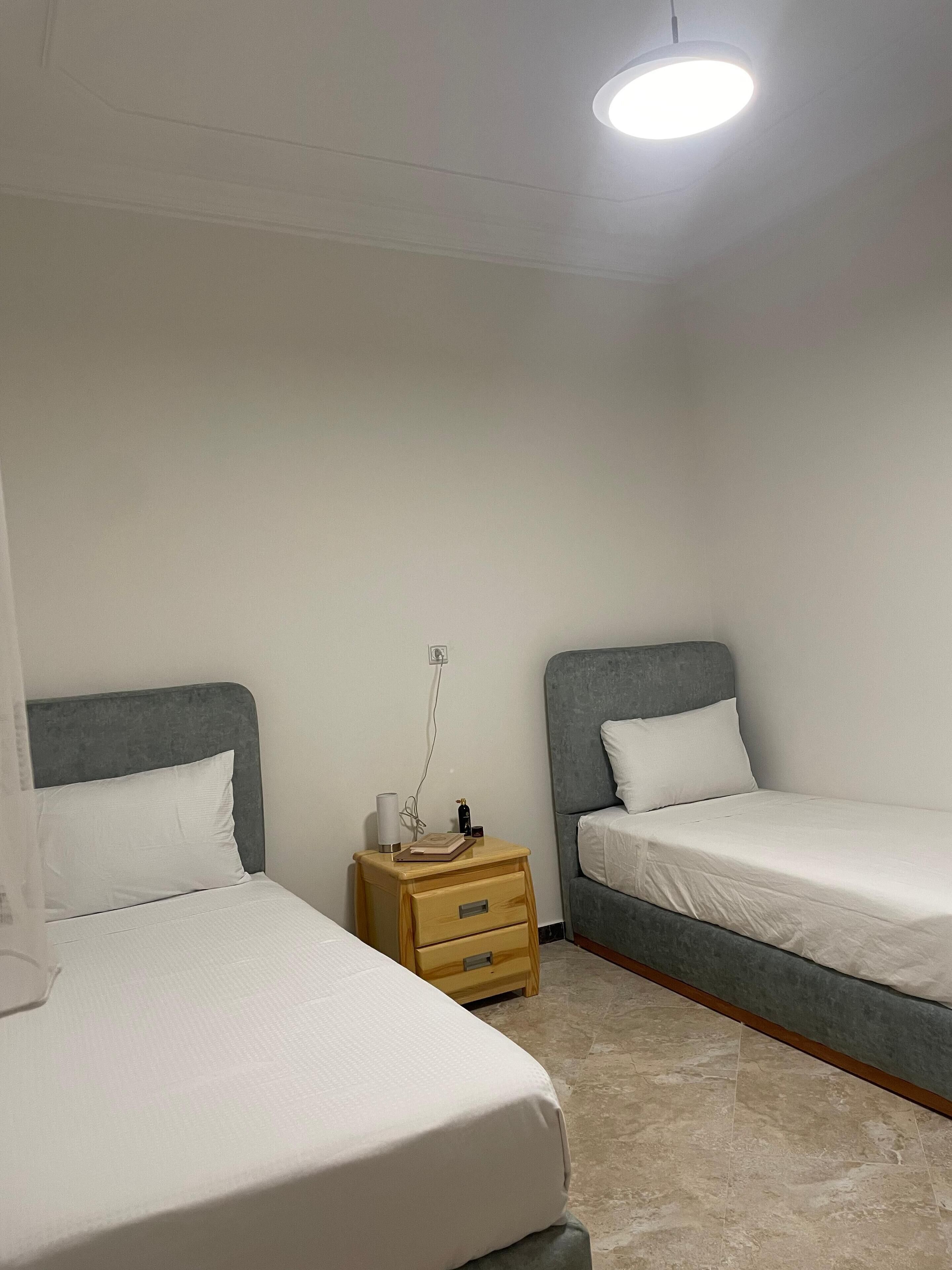 Un dormitorio con dos camas individuales, una mesita de noche de madera con una lámpara y una luz en el techo.