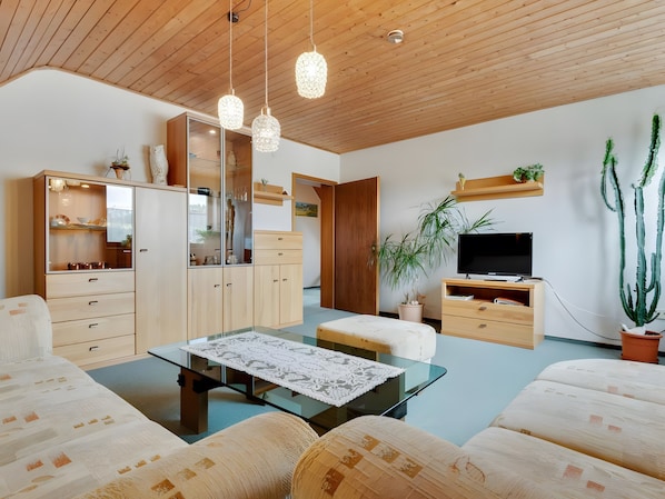 Una sala de estar moderna con una mesa de centro de vidrio, muebles de madera, un televisor y una planta de cactus.