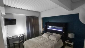 A hotel room with a bed, desk, chair, TV, and a nightstand with a lamp.