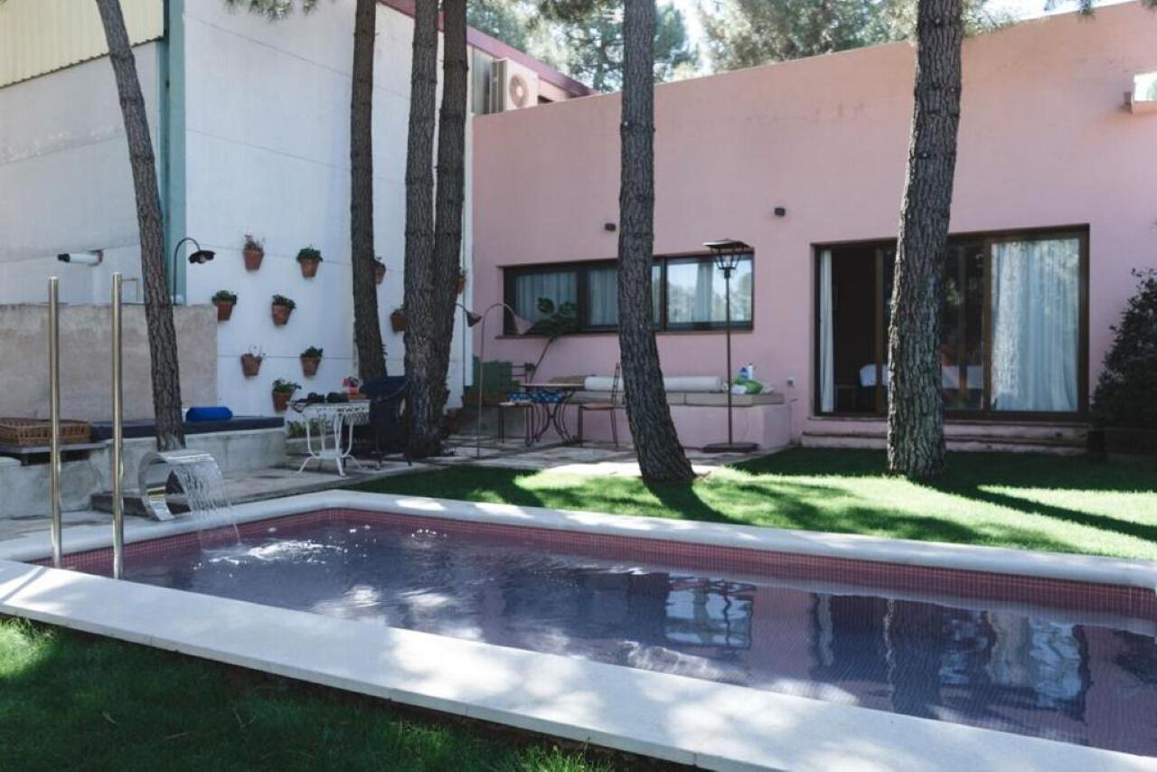 Piscina com deck revestido, rodeada por gramado e árvores. É visível um edifício com fachada rosa e uma janela.