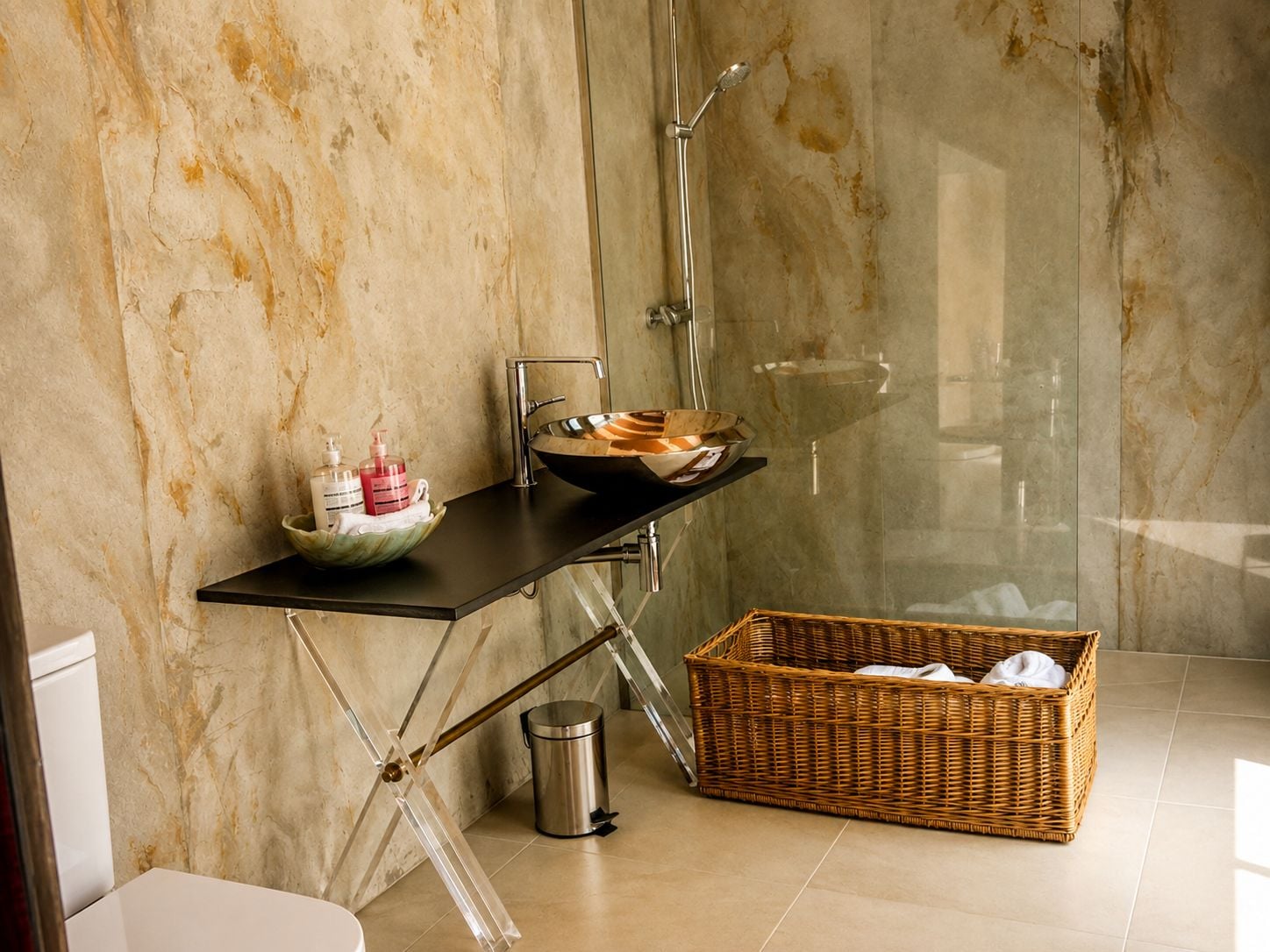 Un baño con pared de mármol, encimera negra, lavabo, ducha, cesta de mimbre y cubo de basura.