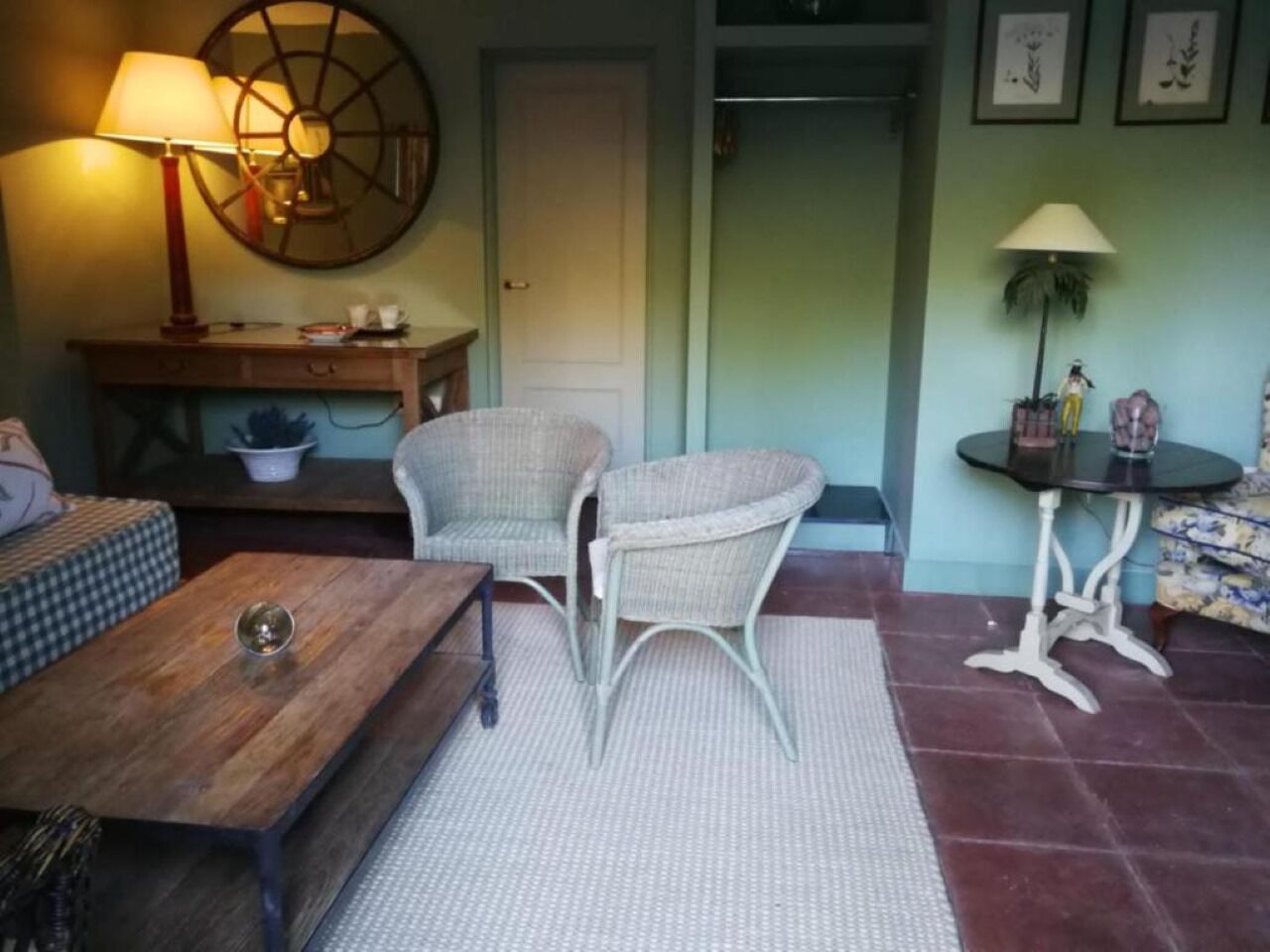 A cozy living room with a wooden coffee table, two wicker chairs, a small round table with a lamp, and a sofa.
