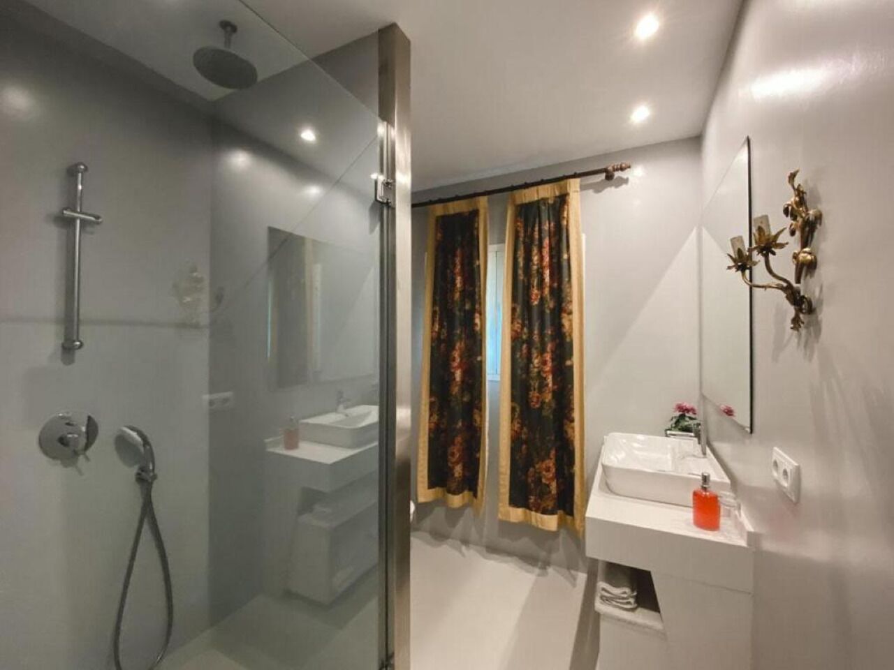 Un baño moderno con ducha de vidrio, lavamanos blanco y espejo con detalles decorativos.