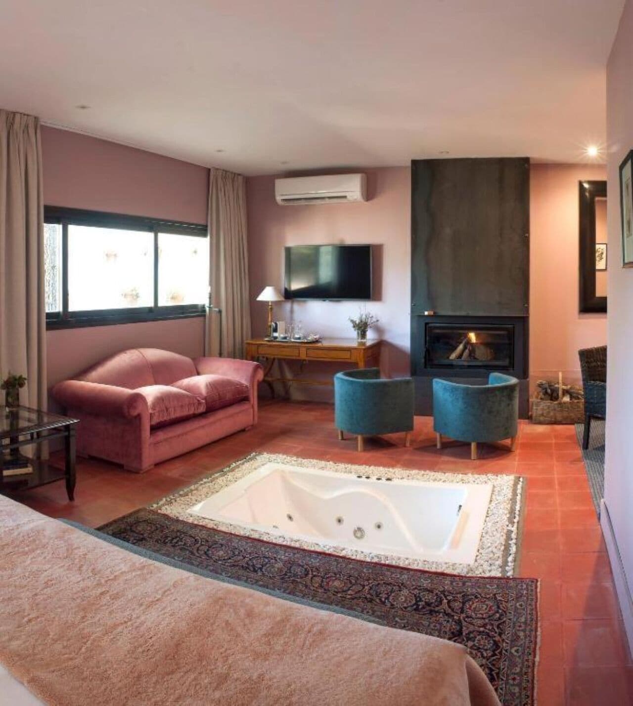 A room with a large bed, a pink sofa, a blue armchair, a small table with a lamp, a flat-screen TV, and a fireplace.
