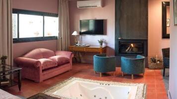 Una habitación con una cama grande, un sofá rosa, un sillón azul, una mesita con una lámpara, un televisor de pantalla plana y una chimenea.