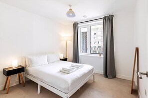 Ein modernes Schlafzimmer mit einem weißen Bett, Nachttischen, einer Lampe und einem Fenster mit Vorhängen.