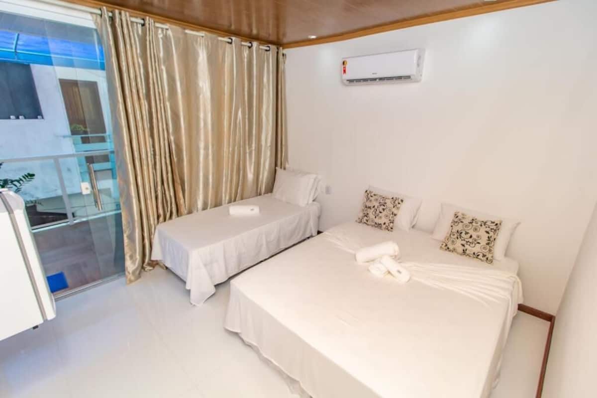 Quarto de hotel com duas camas, uma varanda com cortinas e um aparelho de ar condicionado na parede.