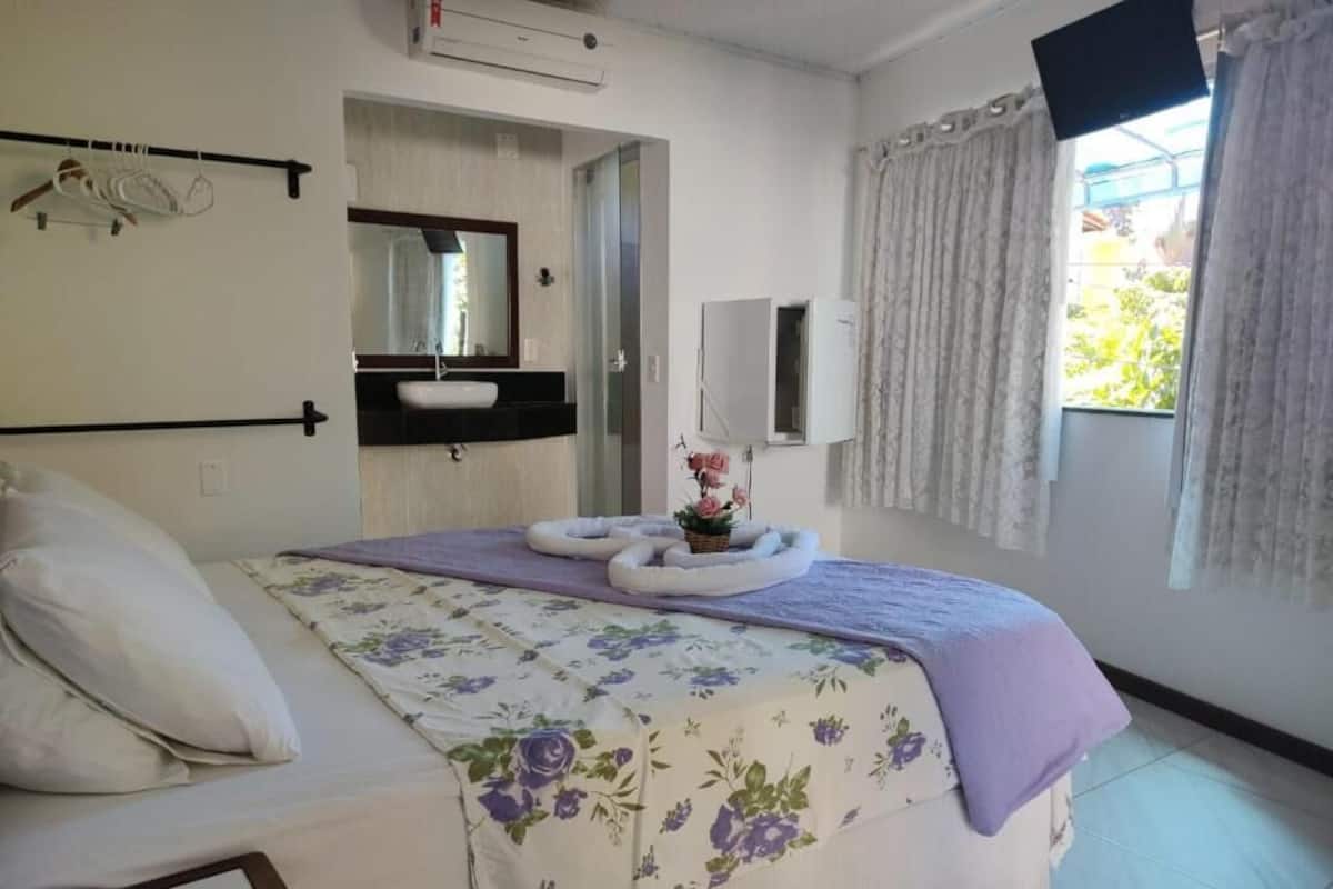 Quarto com cama, mesa de cabeceira, espelho, janela com cortinas e banheiro visível por uma porta aberta.