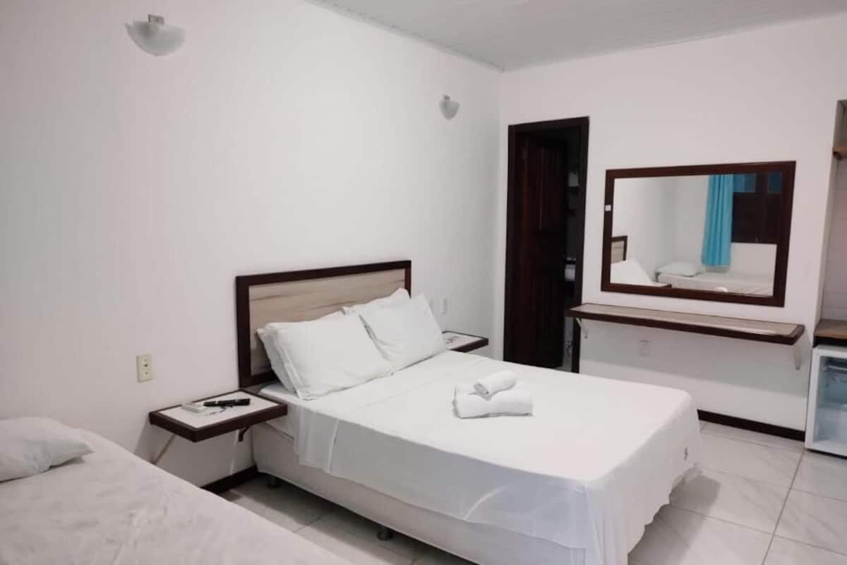 Quarto de hotel com uma cama de solteiro, mesa de cabeceira, espelho e banheiro visível por uma porta aberta.