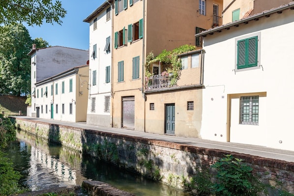 A canal with buildings on one side, featuring balconies with plants, a stone wall, and a clear sky.