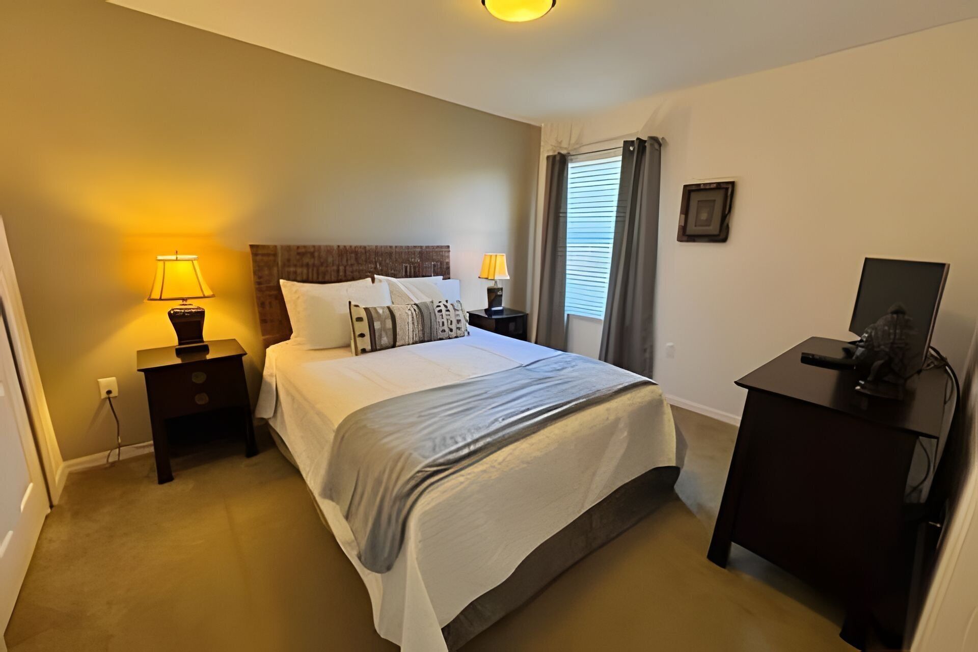 Une chambre à coucher bien rangée, avec un lit, des lampes de chevet, un bureau avec un ordinateur et une fenêtre avec des rideaux.