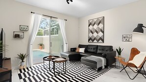 A modern living room with a black and white striped rug, a grey sofa, a coffee table, and a chair. There is a large window with curtains, a potted plant, and a wall-mounted artwork. - Champions Gate, Pool Home near Disney (Kissimmee)