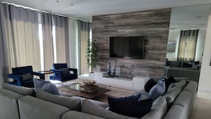 A modern living room with a grey sectional sofa, a blue armchair, a glass coffee table, and a flat-screen TV mounted on a wooden wall. - Renovated Themed Bedrooms, Game Room, Pool (Kissimmee)
