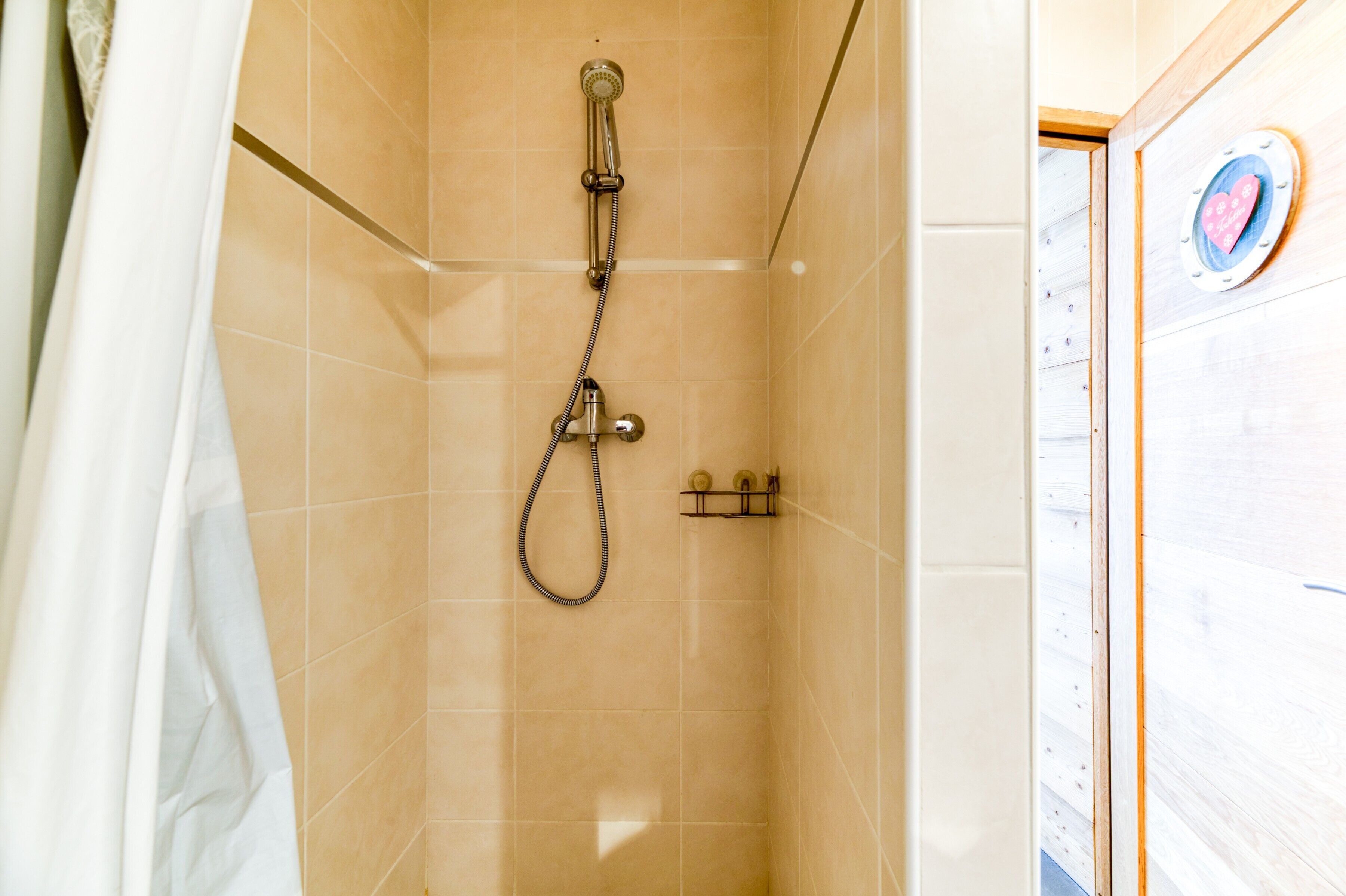 A shower with a handheld showerhead and a wall-mounted faucet, tiled walls, and a wooden door with a heart-shaped decoration.