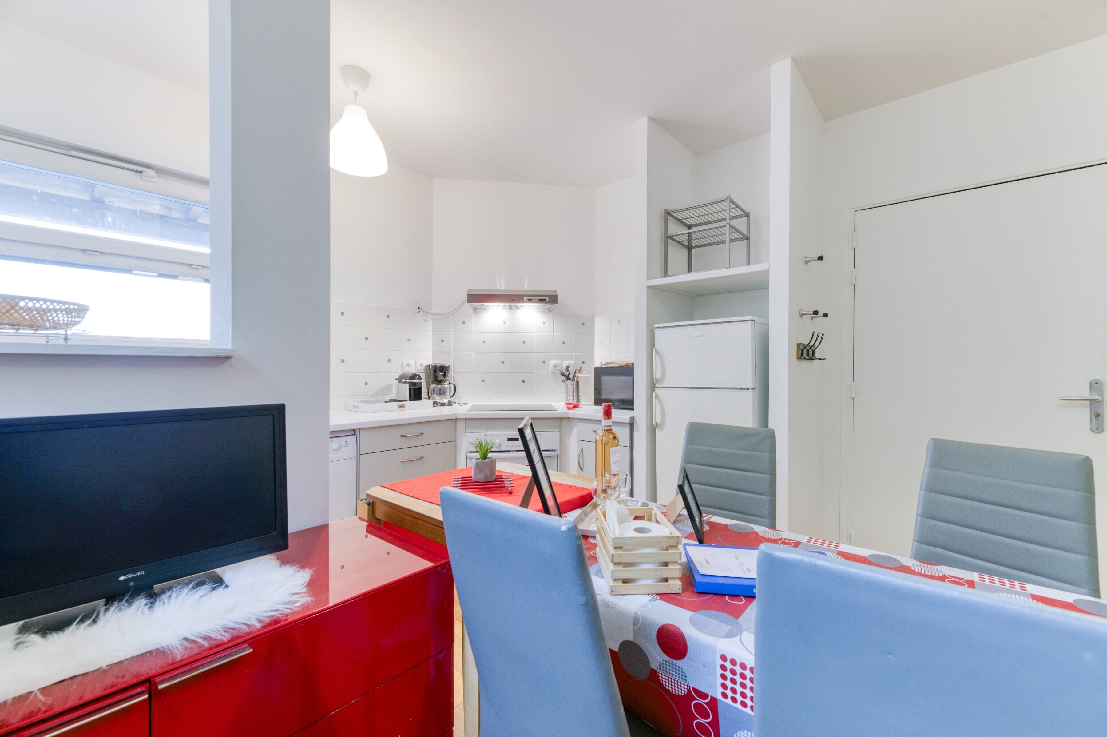 Una cocina compacta con microondas, estufa y armarios, un comedor con mesa y sillas, y un televisor sobre un mueble rojo.