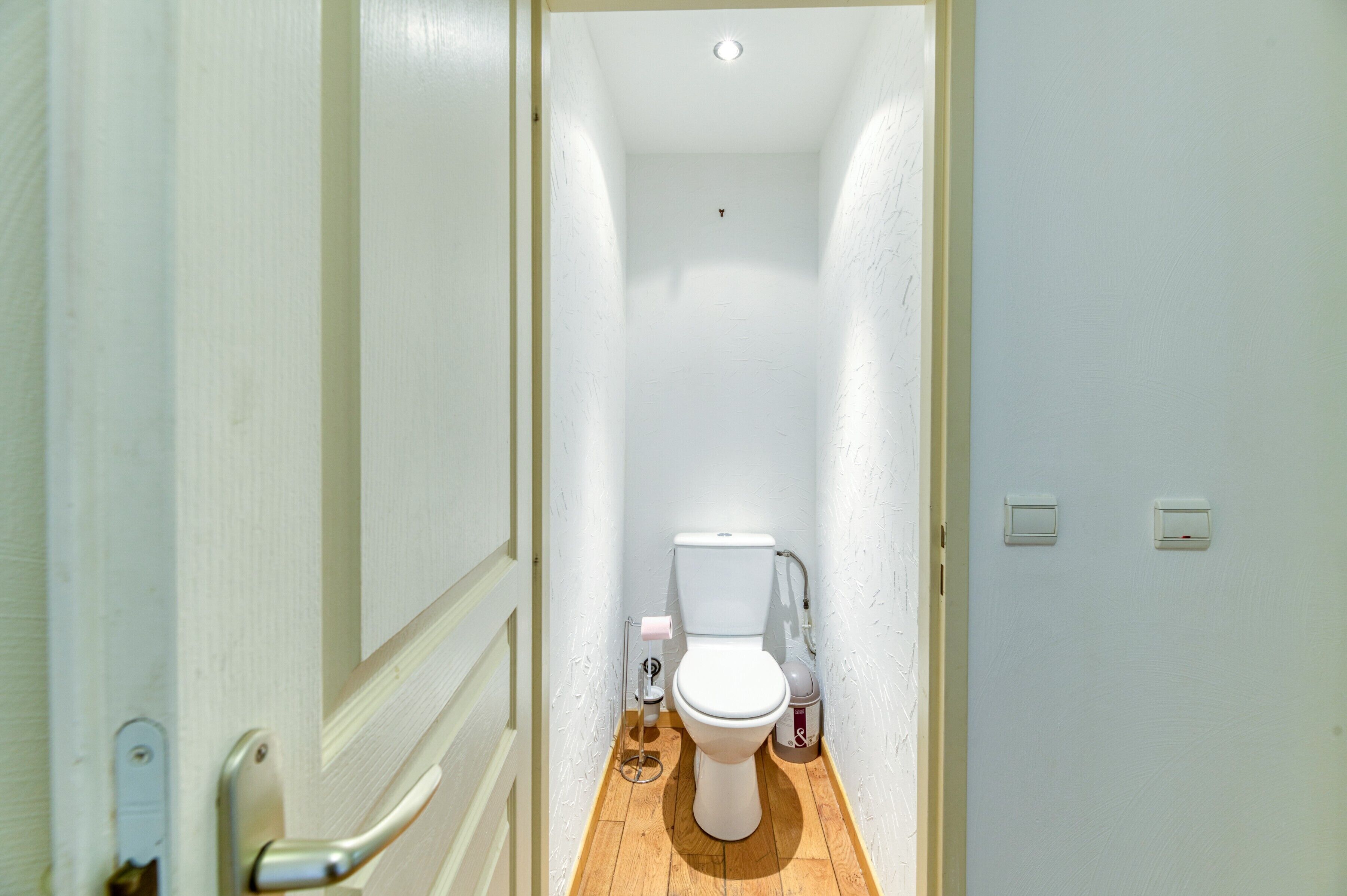 Un couloir étroit mène à des toilettes blanches dont le sol est en bois. Un garde-corps se trouve sur le côté gauche, et deux interrupteurs sont fixés au mur.