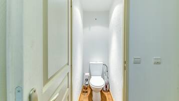 Un couloir étroit mène à des toilettes blanches dont le sol est en bois. Un garde-corps se trouve sur le côté gauche, et deux interrupteurs sont fixés au mur.