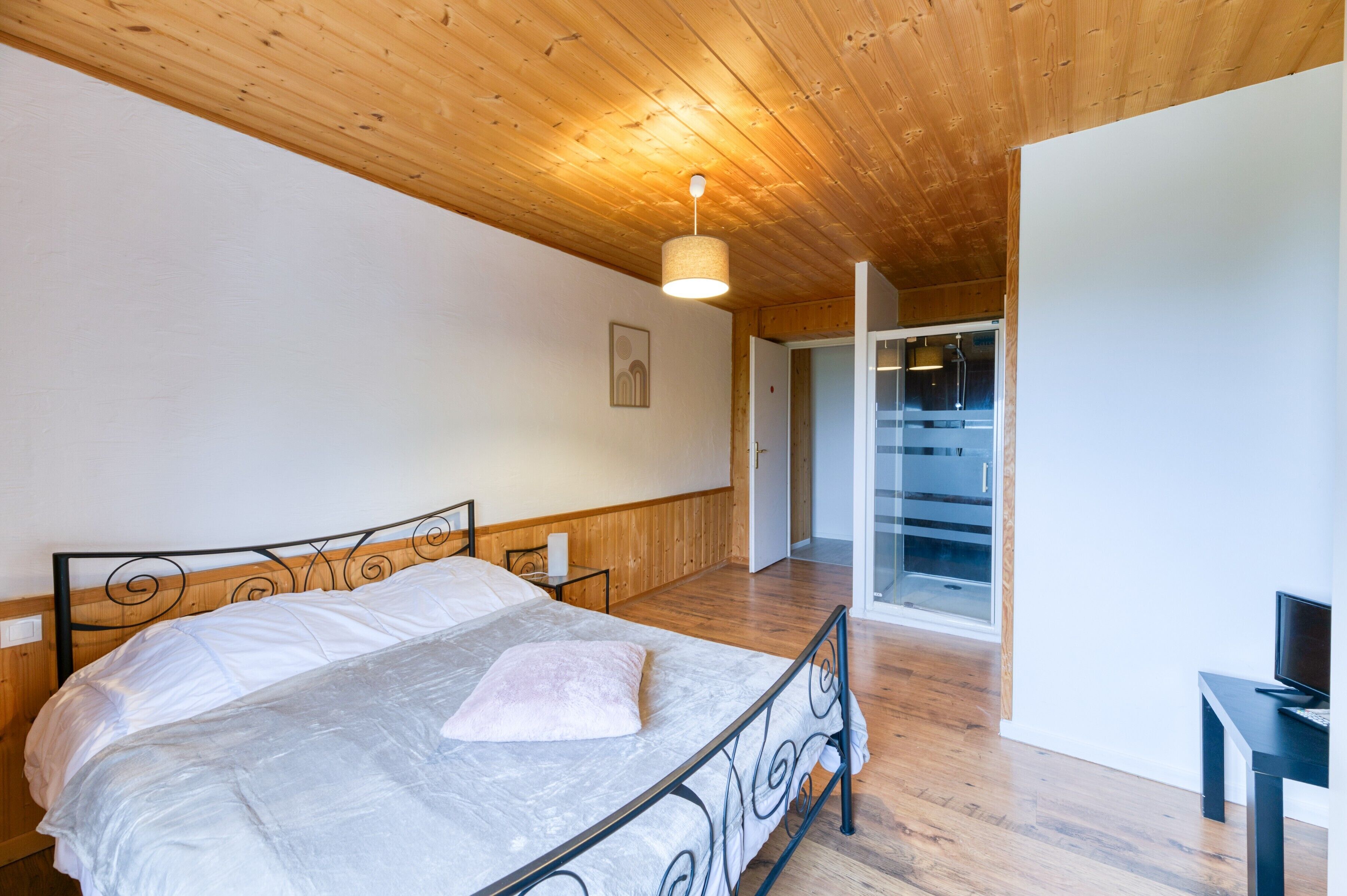 Une chambre à coucher avec un plafond en bois, un lit avec un cadre en métal, une table de chevet et un téléviseur posé sur un meuble.