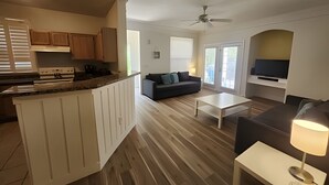 Una sala de estar moderna con sofá, mesa de centro y televisor. Cuenta con una cocina con armarios de madera y encimera. La habitación tiene ventilador de techo y ventanas amplias.