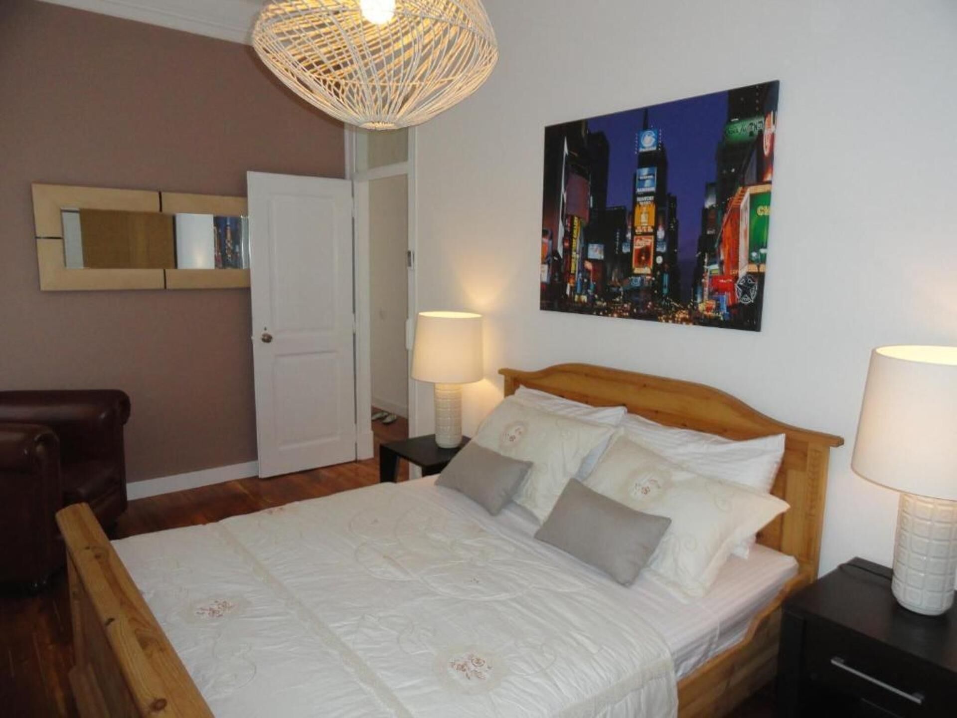 Una camera da letto con un letto in legno, biancheria da letto bianca, un quadro con una veduta cittadina sulla parete, una lampada a soffitto in vimini e una porta sullo sfondo.