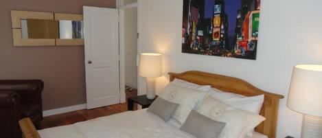 A bedroom with a wooden bed, white bedding, a cityscape painting on the wall, a woven ceiling light, and a door in the background.