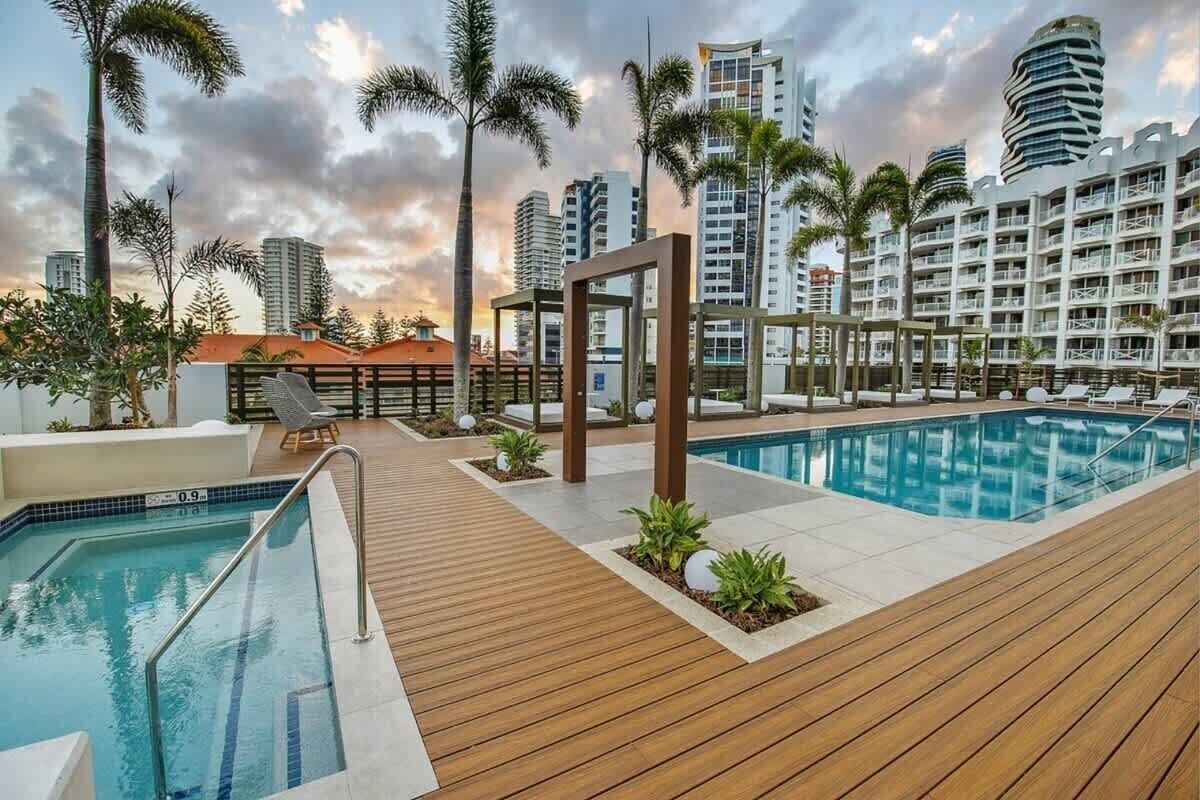 A rooftop pool area with two pools, palm trees, and modern buildings in the background.