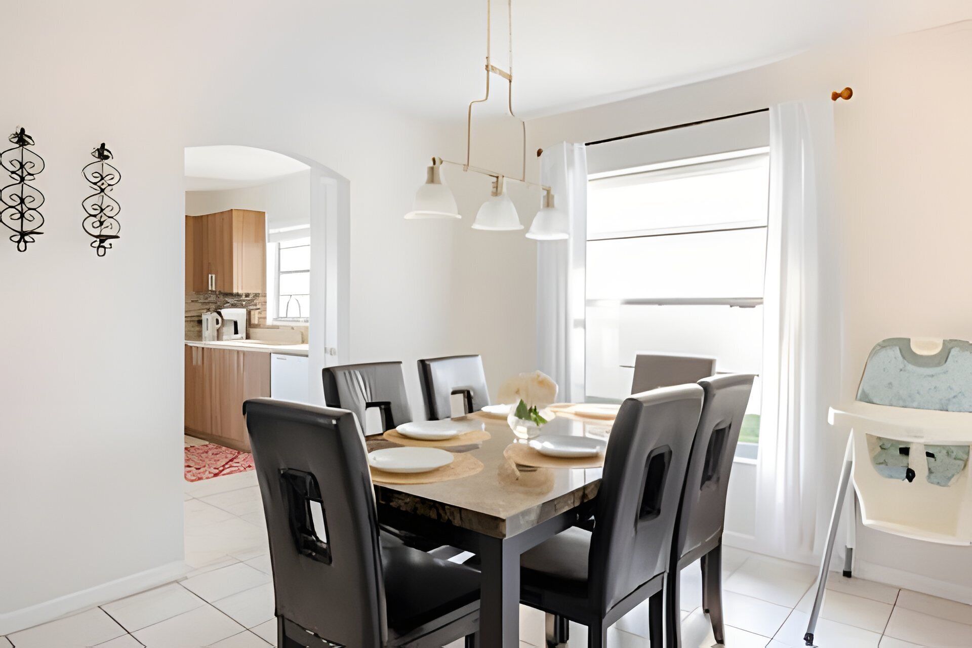 A modern dining area with a wooden table, six chairs, and pendant lights. A kitchen is visible in the background with a window and a view of the outdoors.