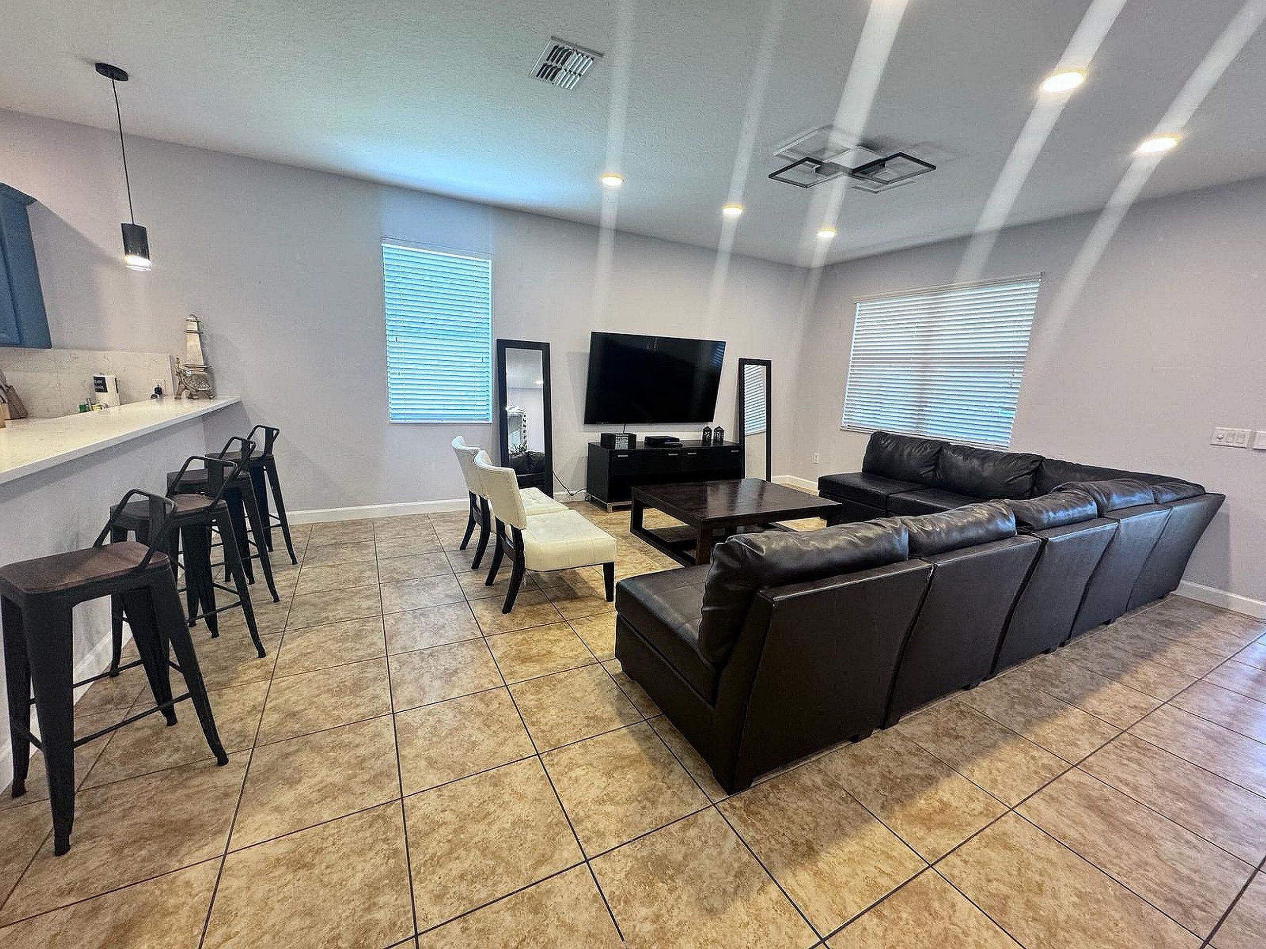 A modern living room with a black leather sofa, a dining area with bar stools, a flat-screen TV, and a kitchen counter.