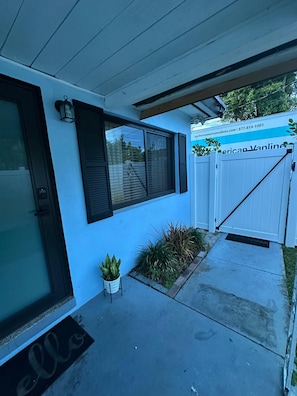 A modern house with a blue exterior, a front door, a window with shutters, a small potted plant, and a gated driveway.