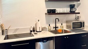 A modern kitchen with a dishwasher, microwave, and a sink with a faucet. There are shelves above the sink holding dishes and a coffee maker. - Charming 1-bedroom house in marvelous Fort Lauderdale with WiFi, AC, Parking (Fort Lauderdale)