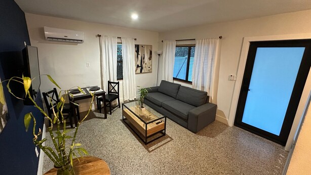 A living room with a sofa, coffee table, dining table, and chairs. There is a plant on the coffee table and a painting on the wall. - Charming 1-bedroom house in marvelous Fort Lauderdale with WiFi, AC, Parking (Fort Lauderdale)
