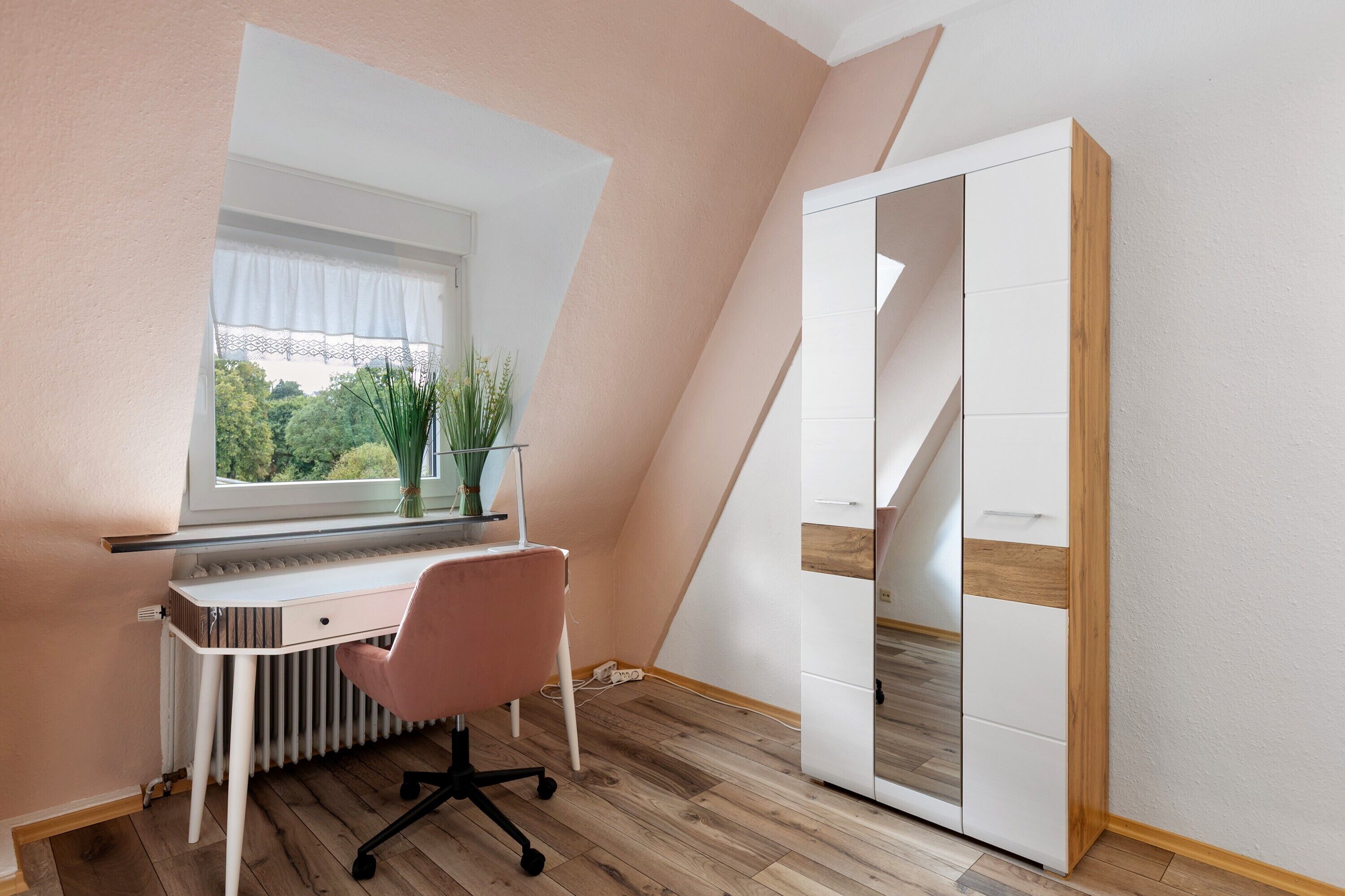 Et værelse med skrå vægge, et skrivebord med stol, en radiator og et vindue med udsigt til grønne omgivelser.