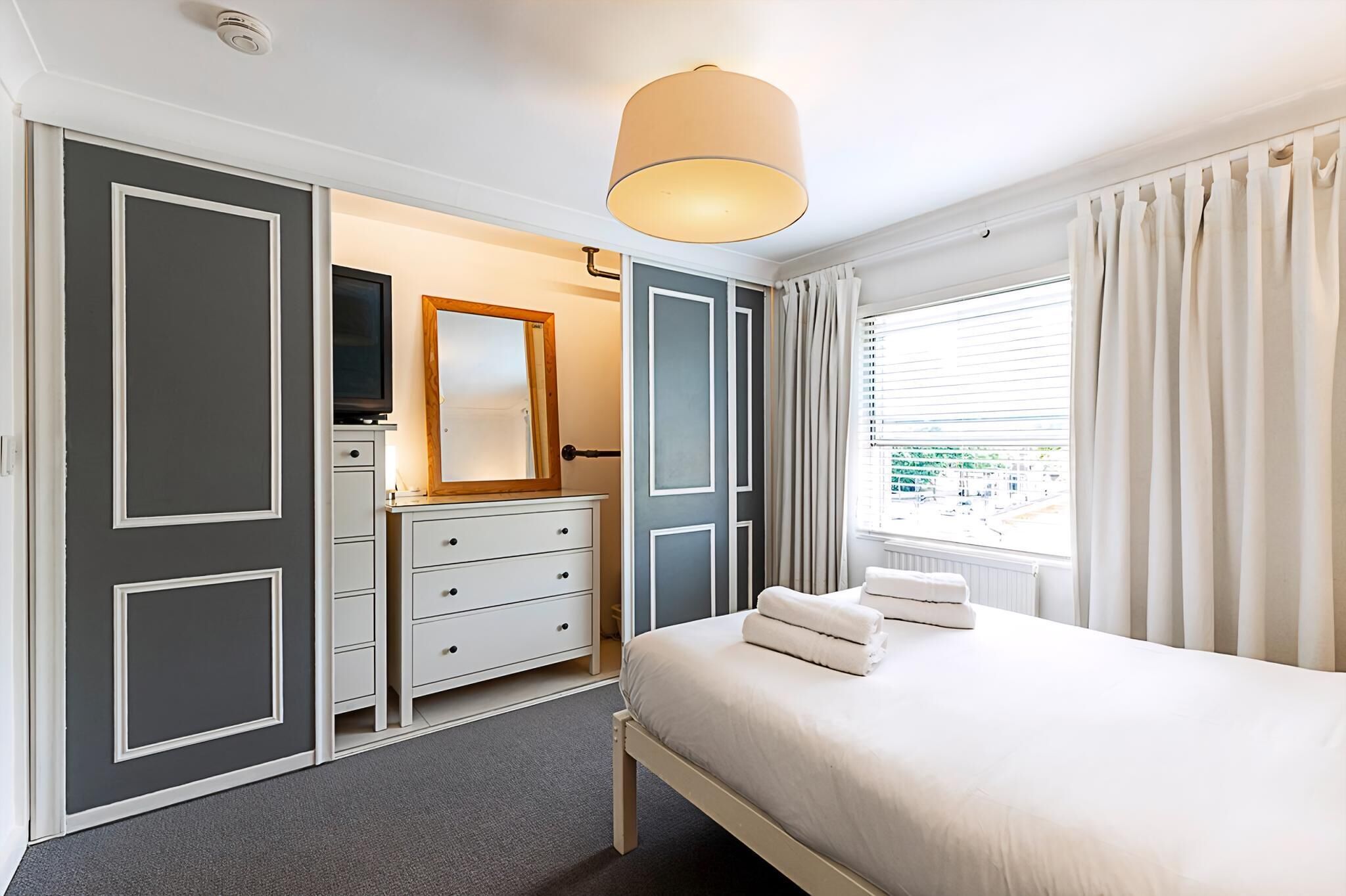 Quarto de hotel com cama, penteadeira, espelho e janela com cortinas.