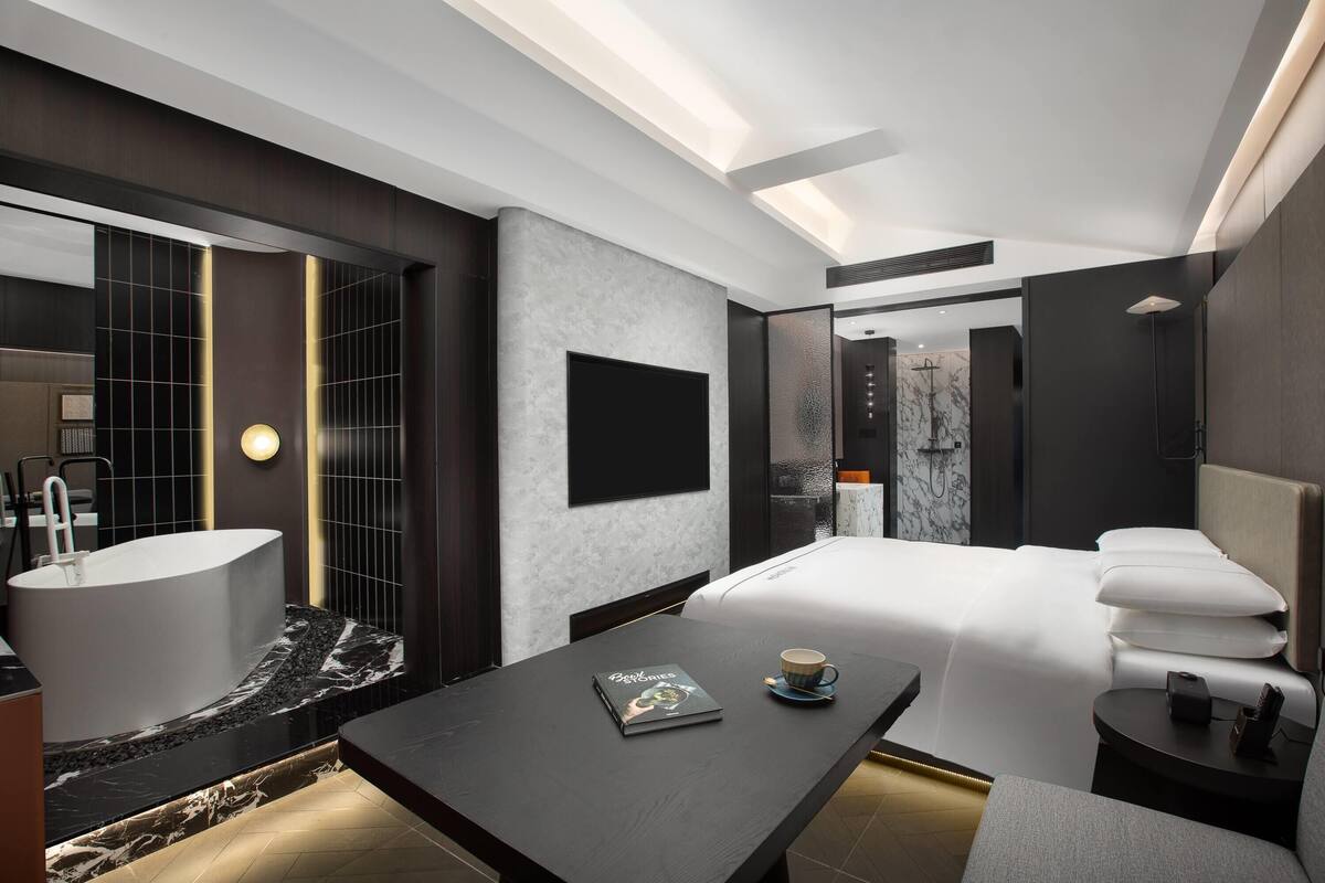 Quarto de hotel moderno com uma cama grande, área de estar com uma mesa, banheira e um espelho amplo.