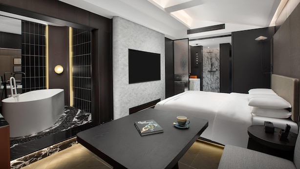 A modern hotel room with a large bed, a sitting area with a table, a bathtub, and a large mirror.