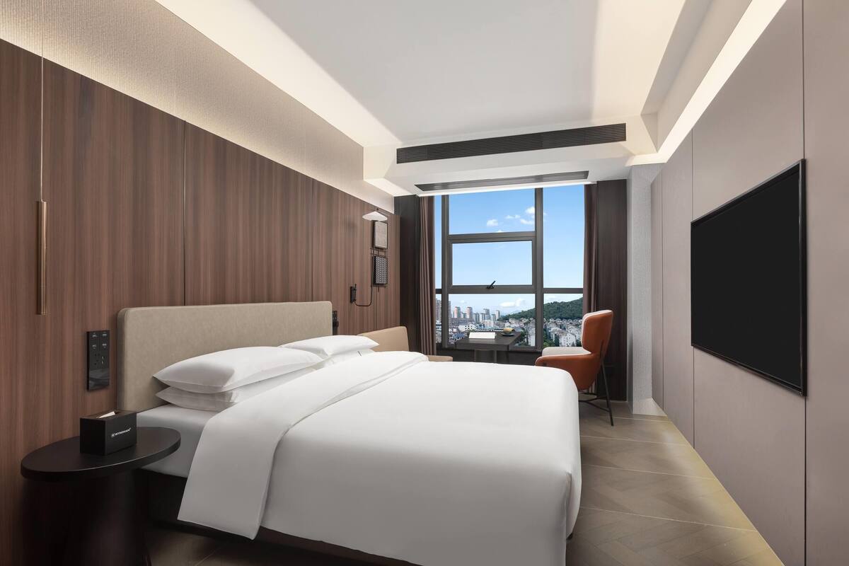 Quarto de hotel moderno com uma cama grande, televisão e vista para a cidade.