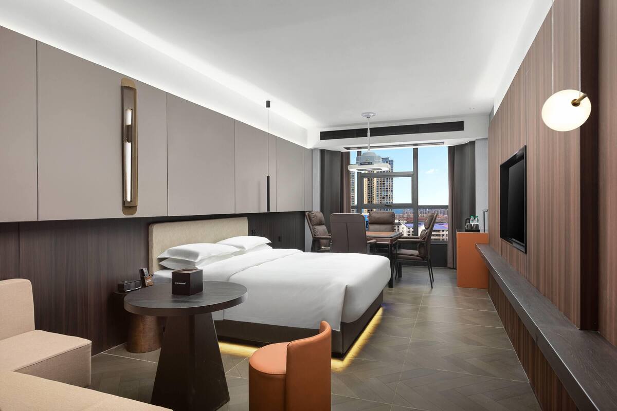 Quarto de hotel moderno com uma cama grande, uma escrivaninha, uma cadeira, televisão e vista para a cidade.