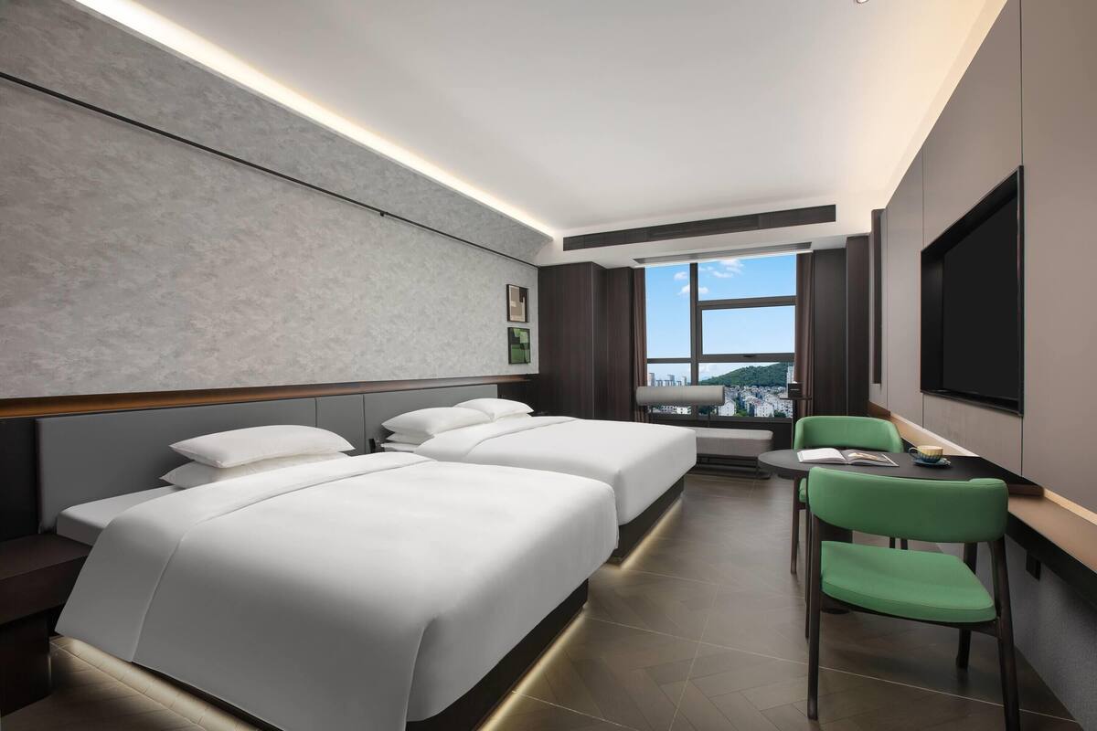 Quarto de hotel moderno com duas camas, uma cadeira verde, uma mesa pequena com um livro e uma xícara, e uma janela grande com vista para a cidade.