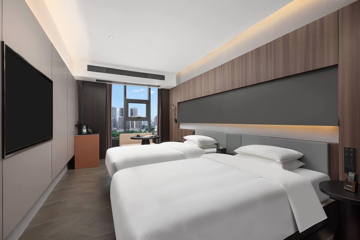 Quarto de hotel moderno com duas camas, uma televisão grande e vista para a cidade.