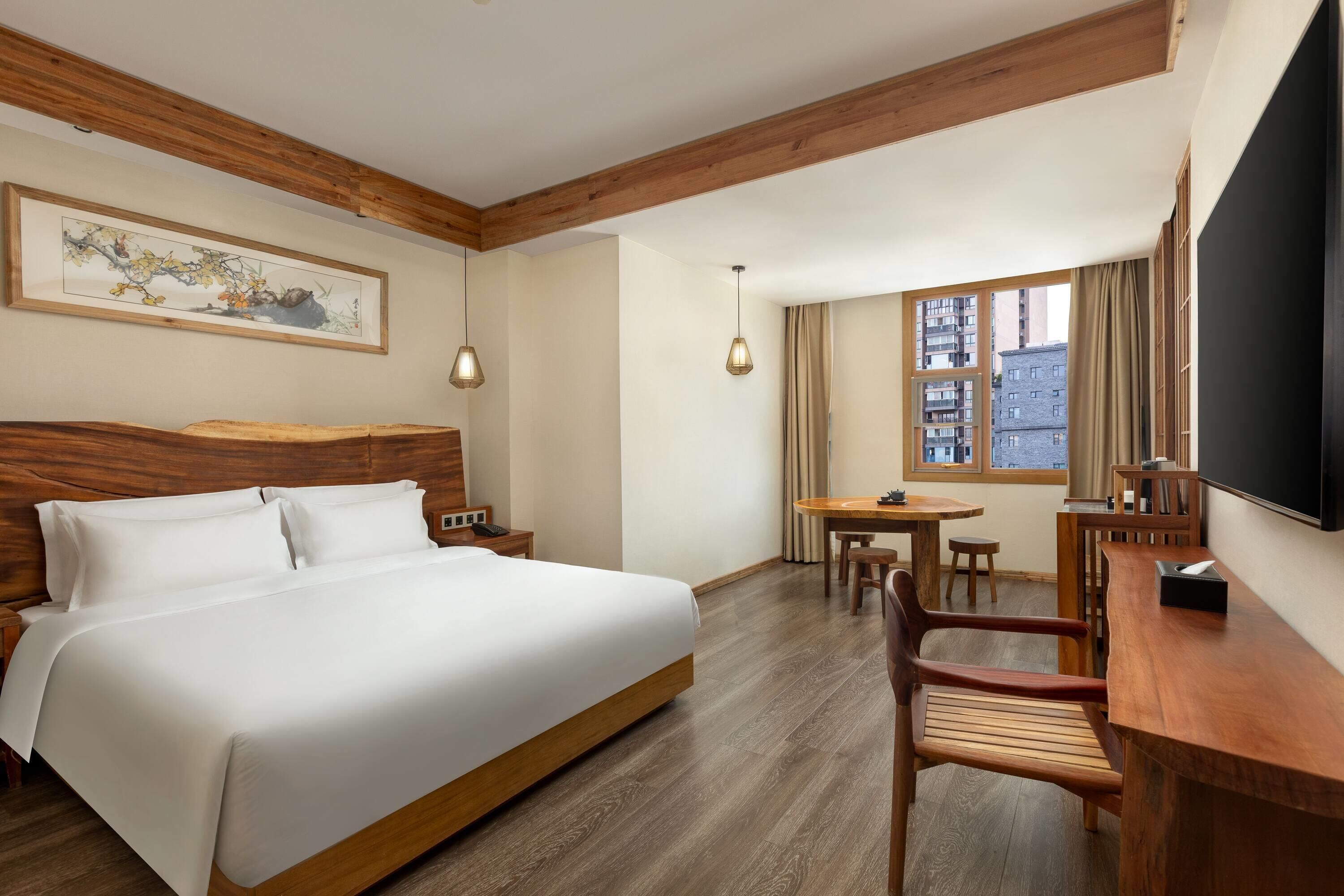 A hotel room with a large bed, wooden headboard, a desk with a chair, a small table, and a TV on the wall.