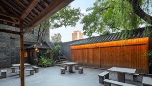 A courtyard with stone benches and tables, a wooden pergola, and a traditional wooden wall with a warm light.