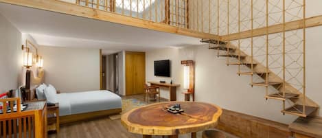 A modern hotel room with a wooden floor, a round wooden dining table, a bed, a desk, a TV, and a staircase with a wooden railing.
