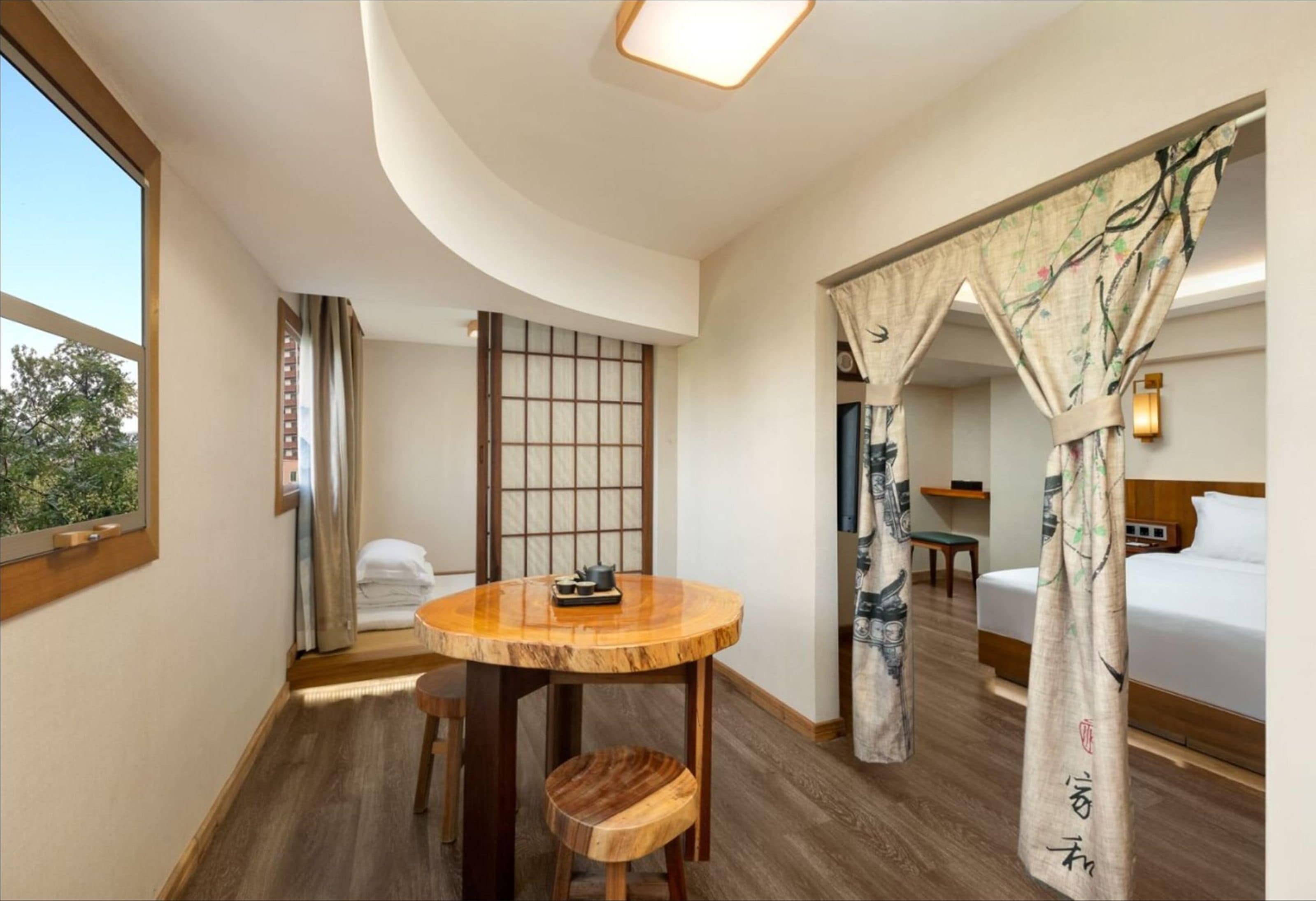 A hotel room with a wooden table, a futon bed, a traditional Japanese shoji screen, and a view of trees outside.